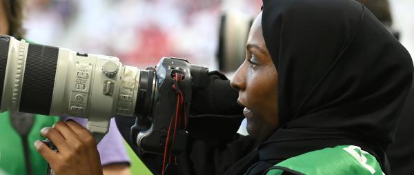 Woman wearing a hijab, holding a camera as she photographs a football match